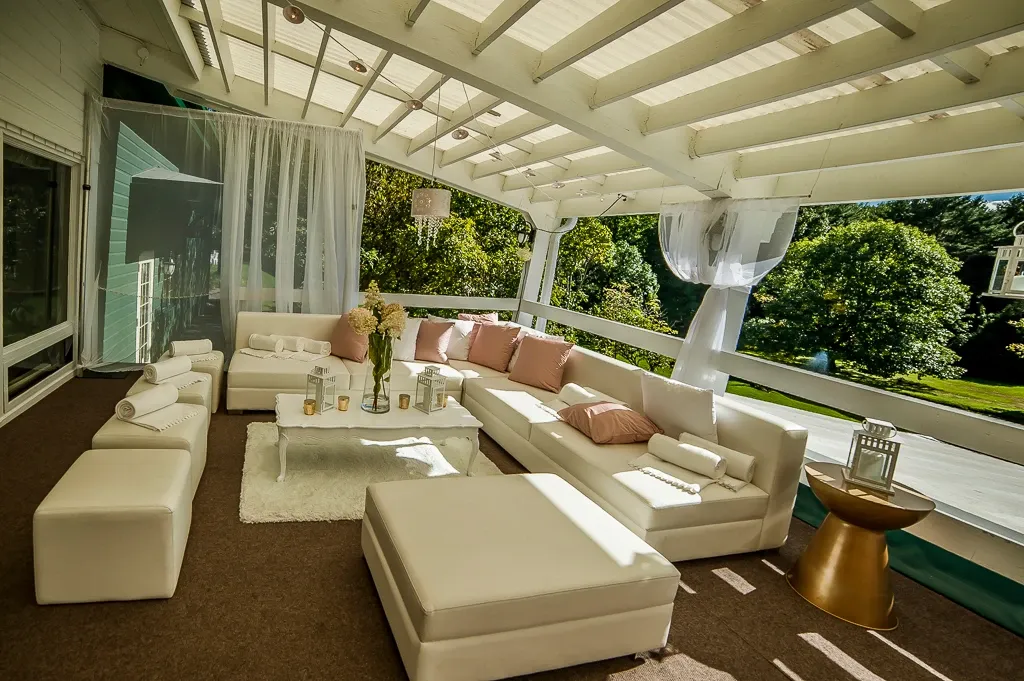 The image showcases a beautifully arranged seating area at La Distinction, a premium wedding venue. This inviting space features elegant white couches adorned with soft pink cushions, creating a cozy and sophisticated ambiance. A large white ottoman and smaller white benches complement the setup, all arranged on a plush white rug. The area is partially enclosed with sheer white drapes, allowing natural light to filter through, adding a romantic glow. Tall vases with delicate flowers and elegant, tall candles enhance the wedding atmosphere. The backdrop of lush green trees provides a serene and picturesque view, ideal for a memorable wedding event.