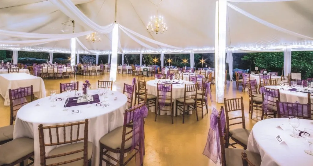 The image showcases an elegant wedding venue by La Distinction, categorized under Wedding Venues on Mariage Elegant, Canada. The spacious setting features round tables draped in white linens, each adorned with delicate purple accents. The chairs are elegantly wrapped with purple sashes, complementing the theme. Soft lighting emanates from chandeliers under a large, white canopy, creating a romantic ambiance. The surrounding greenery adds a natural touch to the sophisticated atmosphere, perfect for a memorable wedding celebration.