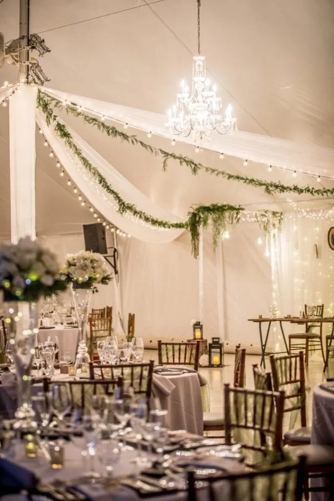 The image showcases an elegant wedding venue setup by La Distinction, categorized under Wedding Venues on Mariage Elegant, Canada. The scene captures a beautifully decorated tent interior, featuring a stunning chandelier casting warm light over the venue. White drapes are artistically arranged with lush greenery, enhancing the romantic ambiance. Dining tables are adorned with elegant tableware and tall floral centerpieces, contributing to a sophisticated atmosphere. The overall setting reflects a perfect blend of elegance and charm, ideal for a memorable wedding celebration.