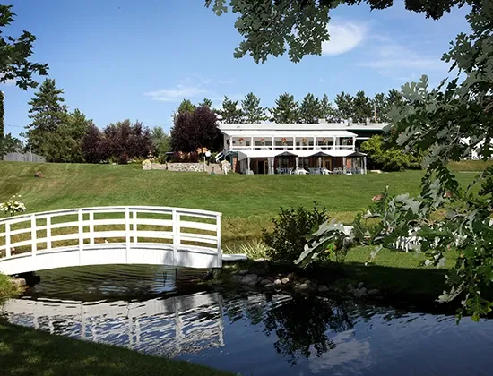 The image showcases La Distinction, a wedding venue in Canada, featured on Mariage Elegant. It depicts a picturesque setting with a lush green lawn leading up to an elegant, two-story venue building surrounded by tall trees. In the foreground, there is a serene pond with a charming white wooden bridge arching over it. The overall scene conveys a tranquil and romantic atmosphere, ideal for hosting memorable wedding ceremonies and receptions.