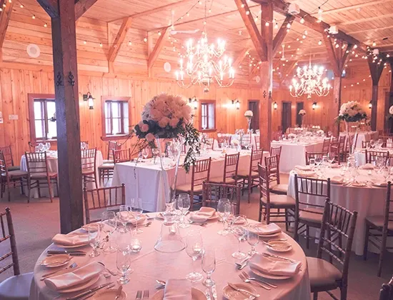 La Distinction, a featured wedding venue on Mariage Elegant in Canada, showcases a beautifully decorated rustic reception hall. The image captures round tables elegantly set with white linens, fine china, and crystal glassware, complemented by floral centerpieces of soft pink and white blooms. The warm wooden interior, adorned with string lights and chandeliers, creates an inviting and romantic ambiance perfect for a wedding celebration.