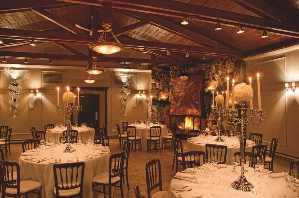 In this image of the wedding venue La Distinction, part of the Mariage Elegant vendor listings in Canada, we see a beautifully arranged reception area. The room features a rustic wooden ceiling with exposed beams and soft, ambient lighting. Circular tables are elegantly set with white tablecloths and ornate candelabras as centerpieces, creating a romantic atmosphere. The walls are tastefully decorated, and there's a striking stone fireplace that adds warmth to the setting. This venue is ideal for couples seeking an intimate and charming location for their wedding celebration.