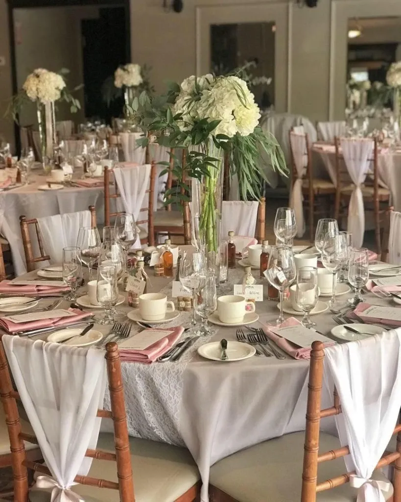 The image depicts an elegantly arranged wedding venue hosted by La Distinction, categorized under Wedding Venues on Mariage Elegant, Canada. The setting includes round tables adorned with white tablecloths and soft pink napkins, complementing the delicate ambiance. Each table is meticulously set with fine china, crystal glassware, and neatly arranged silverware. Centerpieces feature tall vases filled with lush white hydrangeas and greenery, enhancing the sophisticated atmosphere. The wooden chairs are tied with sheer white sashes, adding a touch of elegance to the venue's decor. The overall setting exudes a warm and inviting atmosphere, perfect for a wedding celebration.