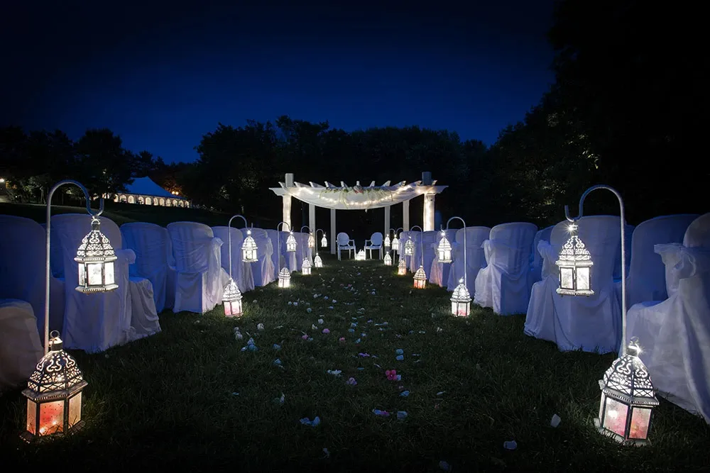 A beautifully illuminated outdoor wedding venue by La Distinction, categorized under Wedding Venues, in Canada. The image showcases a nighttime ceremony setup featuring a white pergola adorned with delicate drapery, framed by a charming aisle lined with ornate lanterns emitting a soft, warm glow. Elegant white chairs draped with covers are arranged on each side of the aisle, which is scattered with pastel flower petals. The serene, tree-lined background enhances the romantic and enchanting atmosphere, making it an ideal setting for a memorable wedding celebration at Mariage Elegant.