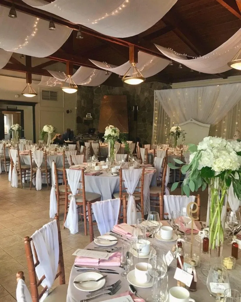 This image showcases an elegant wedding venue by La Distinction, featured in the Wedding Venues category of Mariage Elegant, Canada. The room is beautifully decorated with white draped fabric and string lights hanging from the ceiling, creating a romantic ambiance. Round tables are set with white tablecloths, tall floral centerpieces of white flowers, and delicate glassware, adding to the sophisticated atmosphere. Wooden chairs are adorned with white fabric sashes. The stone wall in the background and the soft lighting enhance the venue's charm, making it an ideal setting for a memorable wedding celebration.