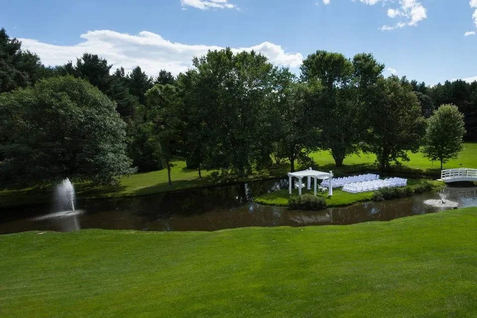La Distinction, a renowned wedding venue in Canada, offers a picturesque outdoor setting for ceremonies. The image showcases a serene landscape with lush green lawns and a tranquil pond. A white aisle with rows of neatly arranged chairs leads to a charming white pergola, providing a focal point for the wedding ceremony. Trees surround the area, contributing to the peaceful ambiance. A white bridge over the pond and a fountain enhance the elegant atmosphere, making it an ideal location for a memorable wedding event.