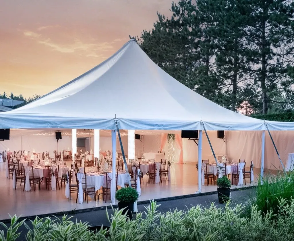 The image shows an elegant outdoor wedding venue by La Distinction, part of the Wedding Venues category on Mariage Elegant, Canada. A large, white tent is set up in a serene, natural setting with lush greenery and tall trees in the background. Inside the tent, round tables are beautifully arranged with white tablecloths and wooden chairs. The tables are elegantly set with glassware and cutlery, creating an inviting atmosphere for a wedding reception. The soft lighting under the tent adds a warm glow, enhancing the romantic ambiance as the sun sets in the sky.
