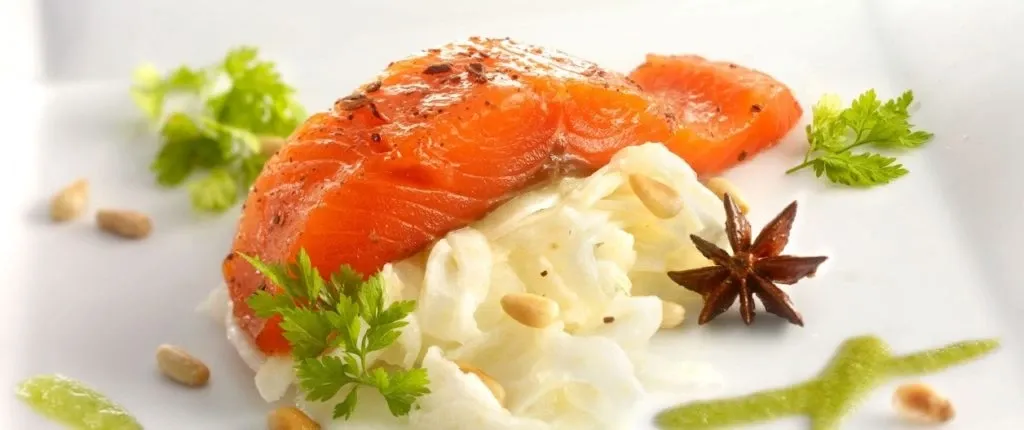 The image showcases a beautifully plated dish featuring a piece of cured salmon atop a bed of creamy, finely shredded cabbage or similar vegetable. The presentation is enhanced with garnishes such as fresh herbs and pine nuts, adding texture and color. The vibrant orange of the salmon contrasts with the white base, and there are accents of sauce and spices thoughtfully arranged on the elegant white plate. This culinary creation emphasizes the exquisite dining options available at La Distinction, a distinguished wedding venue in Canada, perfect for couples seeking a refined and elegant celebration.