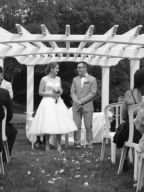 A bride and groom stand smiling under a white pergola at La Distinction, a renowned wedding venue in Canada. The bride is wearing a classic white dress and holding a bouquet, while the groom is dressed in a light-colored suit and bow tie. They are surrounded by seated guests in a picturesque outdoor setting with lush greenery in the background, perfect for a romantic garden wedding. The image captures the joyful ambiance and elegant arrangement at this popular wedding venue.