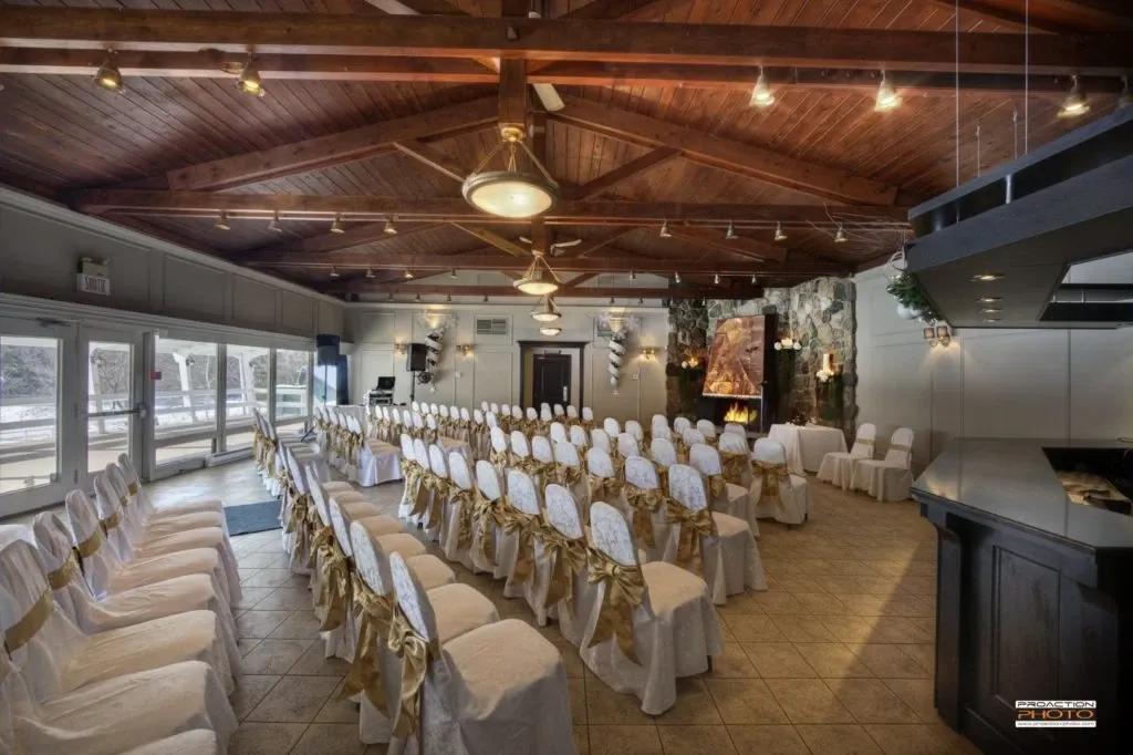 The image shows an elegantly arranged wedding venue, La Distinction, categorized under Wedding Venues on Mariage Elegant, Canada. The space features rows of white chairs adorned with gold bows, set up in a spacious room with a high wooden ceiling and large windows that let in natural light. At the back of the room, a stone fireplace adds a warm ambiance, while decorative elements and indoor plants complement the sophisticated setting. This venue offers a charming and intimate atmosphere, perfect for a memorable wedding ceremony.