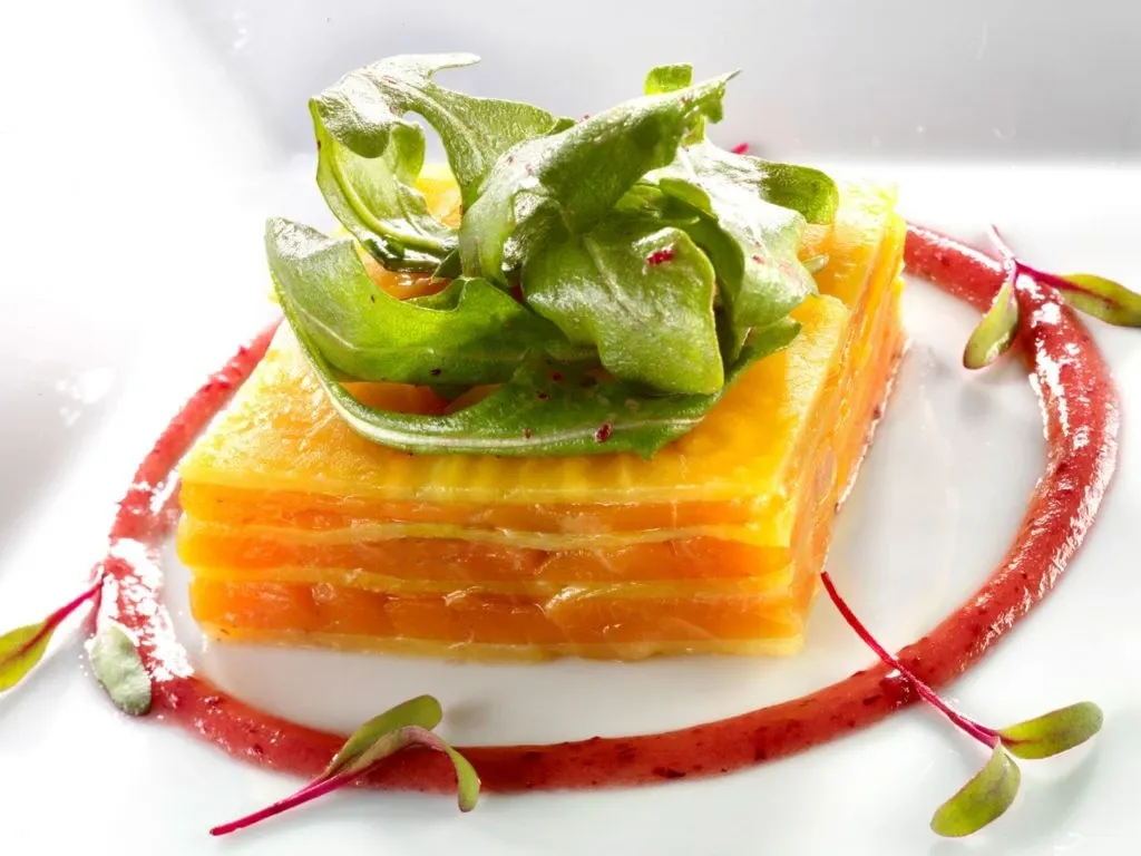 A beautifully plated dish featured at La Distinction, a renowned wedding venue in Canada, specializing in elegant wedding experiences. The culinary presentation includes a stack of thinly sliced yellow and orange vegetables, artfully arranged into a square. It is garnished with fresh green leaves on top and surrounded by a decorative swirl of vibrant red sauce. This exquisite dish reflects the venue's dedication to providing gourmet dining options for weddings and special events.