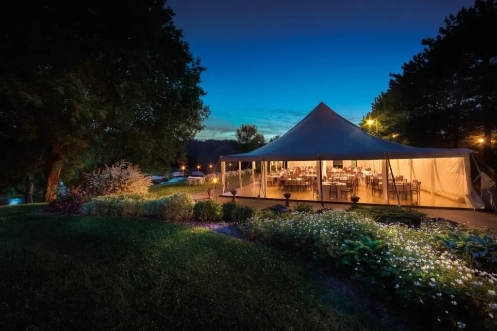 The image features an elegant outdoor wedding venue by La Distinction, highlighting their expertise in providing stunning settings for special occasions. Set against a picturesque evening backdrop, a large, illuminated tent is seen surrounded by lush greenery and well-maintained gardens. The interior of the tent is beautifully decorated, with tables elegantly arranged for a wedding reception, showcasing a warm and inviting atmosphere. Soft lighting enhances the romantic ambiance, making it a perfect choice for weddings hosted at La Distinction in Canada.