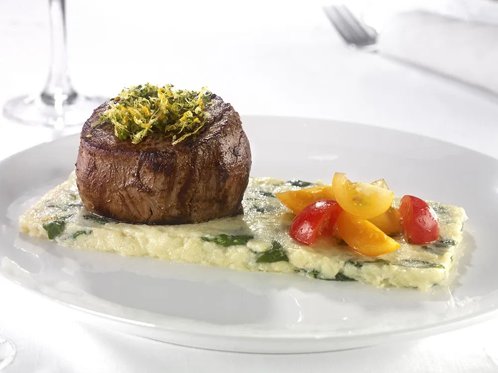 A beautifully presented gourmet dish featuring a perfectly seared filet mignon topped with a garnish of finely chopped herbs and lemon zest rests on a white plate. The steak is paired with a serving of creamy mashed potatoes mixed with asparagus pieces and lies beneath a colorful medley of red and yellow cherry tomato halves. This elegant culinary creation exemplifies the exquisite dining options available at La Distinction, a top-tier wedding venue listed on Mariage Elegant, Canada, perfect for couples seeking a sophisticated and memorable wedding experience.