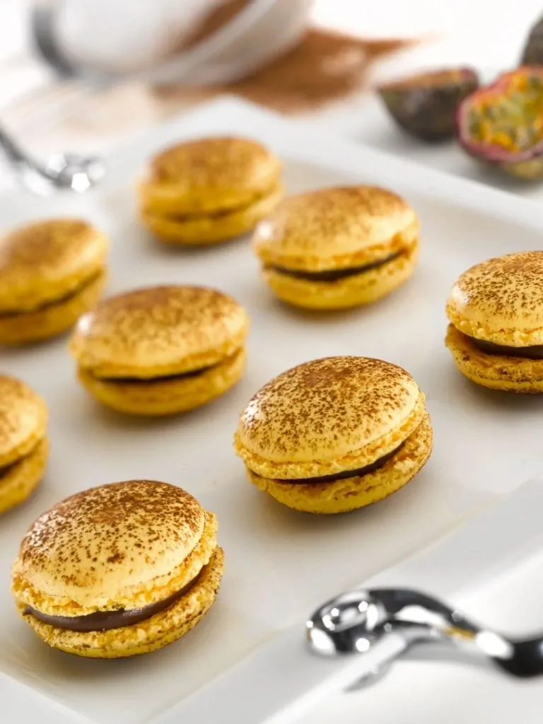 This image features a display of exquisite macarons with a golden-brown dusting, artfully arranged on a pristine white tray. These elegant desserts are positioned in neat rows, showcasing their perfect round shape and creamy filling. The photo conveys a sense of refinement that aligns with the high standards of La Distinction, a premier wedding venue listed on Mariage Elegant in Canada. In the background, there is a glimpse of sliced passion fruit, adding a touch of exotic flair and highlighting the attention to detail expected in luxury wedding catering. This image reflects the sophistication and culinary artistry typical of top-tier wedding venues.