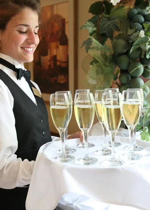 A server from La Distinction, categorized under Wedding Venues, elegantly presents a tray of champagne glasses, perfect for a wedding celebration. The server is dressed in formal attire with a vest and bow tie, holding a white-cloth-covered tray featuring six flutes filled with sparkling champagne. In the background, there is lush green foliage adding a touch of natural decor, ideal for a sophisticated wedding atmosphere at Mariage Elegant in Canada.