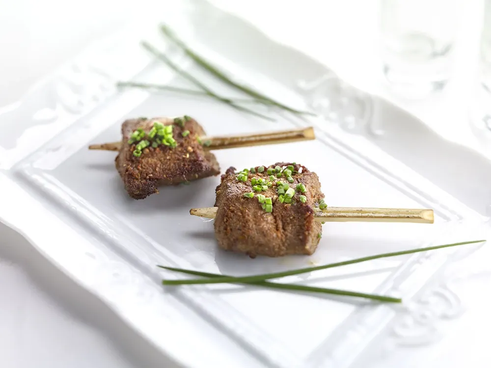 The image depicts a beautifully arranged dish prepared by La Distinction, showcasing their expertise as a wedding venue. Two elegantly plated skewers of seasoned and grilled meat are garnished with finely chopped green herbs, presented on a decorative white rectangular platter. Delicate lines of fresh chives are artistically placed beside the skewers, enhancing the visual appeal. This presentation reflects the attention to detail and gourmet quality offered by La Distinction, a premium vendor in the Wedding Venues category for Mariage Elegant in Canada.