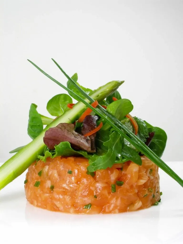 A beautifully plated dish of salmon tartare garnished with fresh greens, carrot shreds, and asparagus spears is artfully presented on a white background. This elegant culinary creation, associated with La Distinction, falls under the Wedding Venues category on Mariage Elegant, Canada. The visually appealing presentation highlights the venue's commitment to offering exquisite dining experiences for weddings.