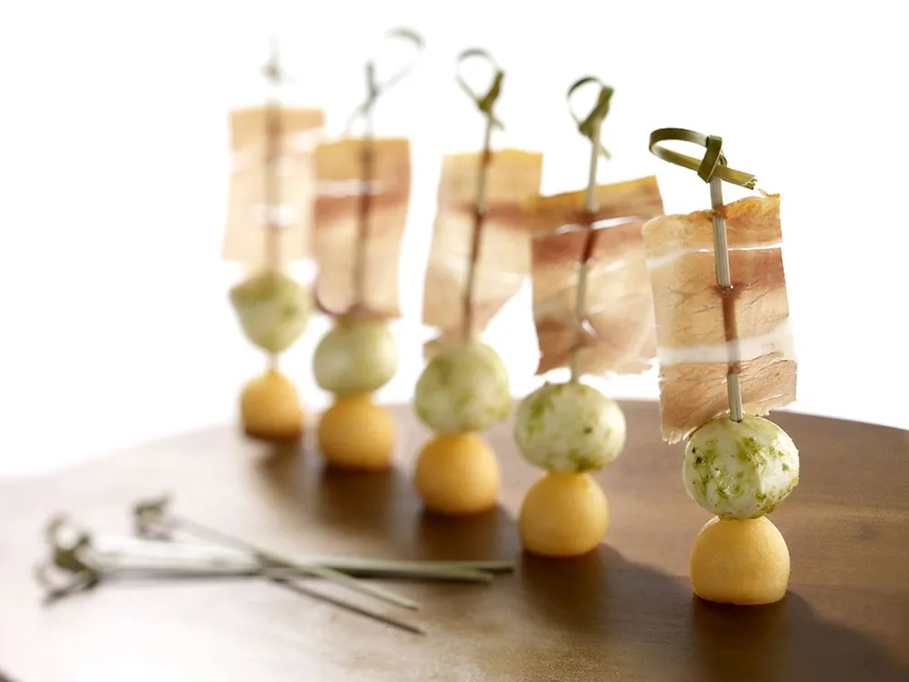 A beautifully arranged display of gourmet appetizers on skewers, each featuring delicate slices of cured meat, soft cheese balls with herbs, and small bread rounds, elegantly presented on a wooden board. This culinary presentation is part of La Distinction's offerings in the Wedding Venues category for Mariage Elegant, Canada, showcasing their attention to detail and high-quality catering services.