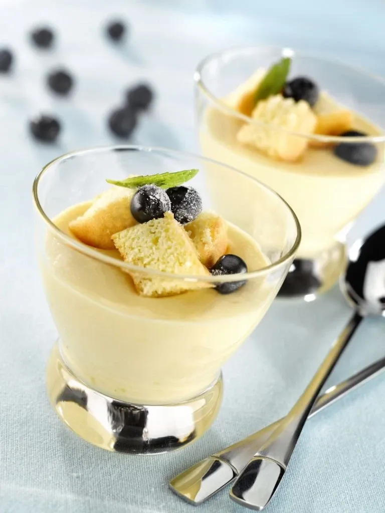 This image showcases a beautifully arranged dessert, featuring two elegant glass cups filled with creamy custard at La Distinction, a distinguished wedding venue in Canada. Each cup is topped with a garnish of fresh blueberries, a sprig of mint, and small cubes of cake, adding a touch of sophistication. The dessert is set on a light blue tablecloth, accompanied by metallic dessert spoons, creating a refined presentation perfect for weddings and special events. La Distinction provides exquisite culinary delights as part of their exceptional wedding venue offerings.