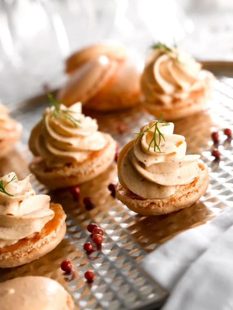 The image showcases an elegant display of gourmet macarons topped with swirls of creamy filling, meticulously arranged on a textured silver tray. Small sprigs of herbs garnish the macarons, adding a touch of sophistication. Pink peppercorns are scattered around, enhancing the luxurious presentation. This culinary detail is from La Distinction, falling under the Wedding Venues category, highlighting its refined taste and attention to detail, ideal for upscale wedding receptions associated with Mariage Elegant in Canada.
