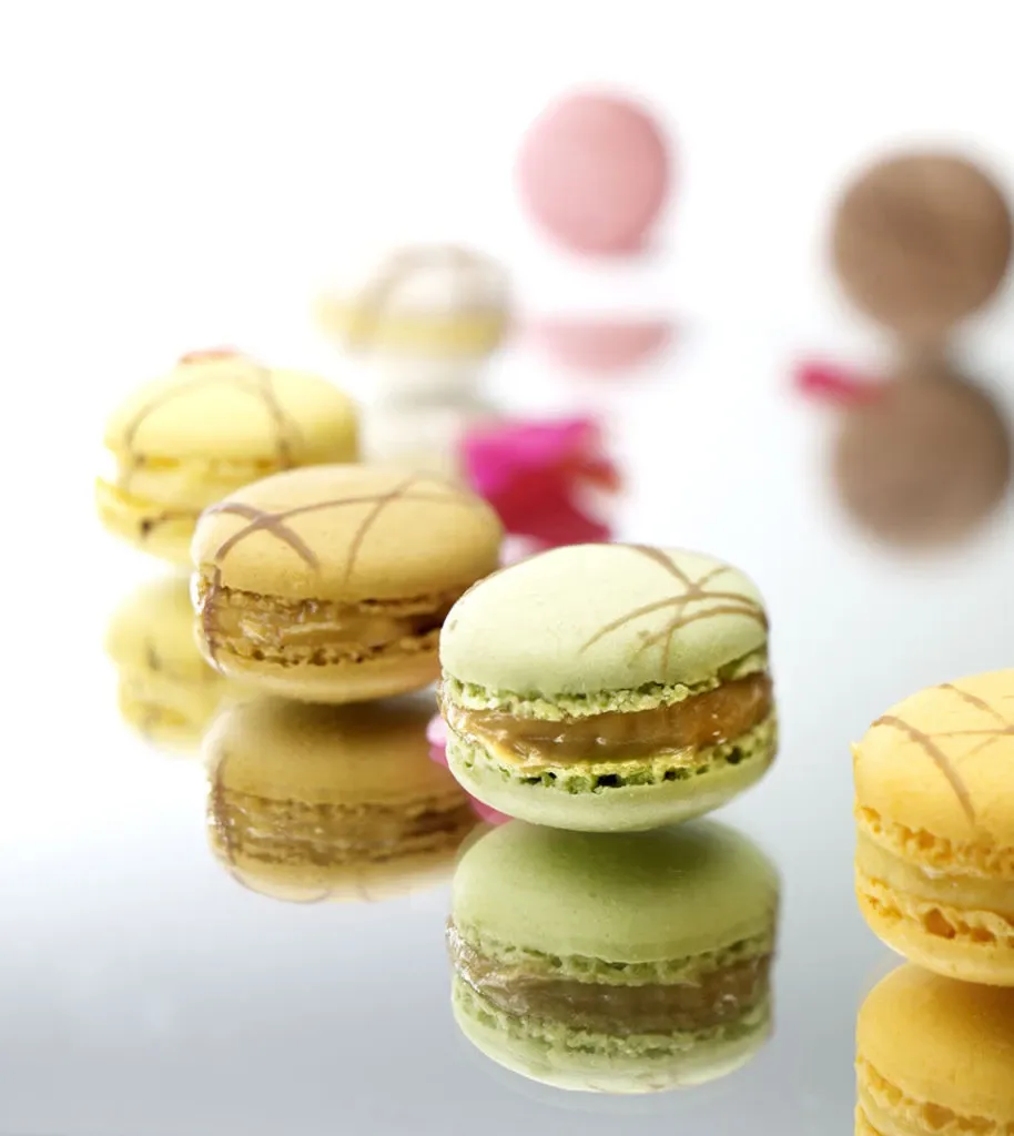 The image shows an elegant presentation of colorful macarons in pastel shades, including yellow, green, and pink, displayed on a glossy surface. The arrangement suggests a delicate and sophisticated wedding dessert option provided by La Distinction, categorized under Wedding Venues on Mariage Elegant, Canada. The reflection on the surface adds a touch of luxury, making it a fitting choice for a stylish wedding reception setting.