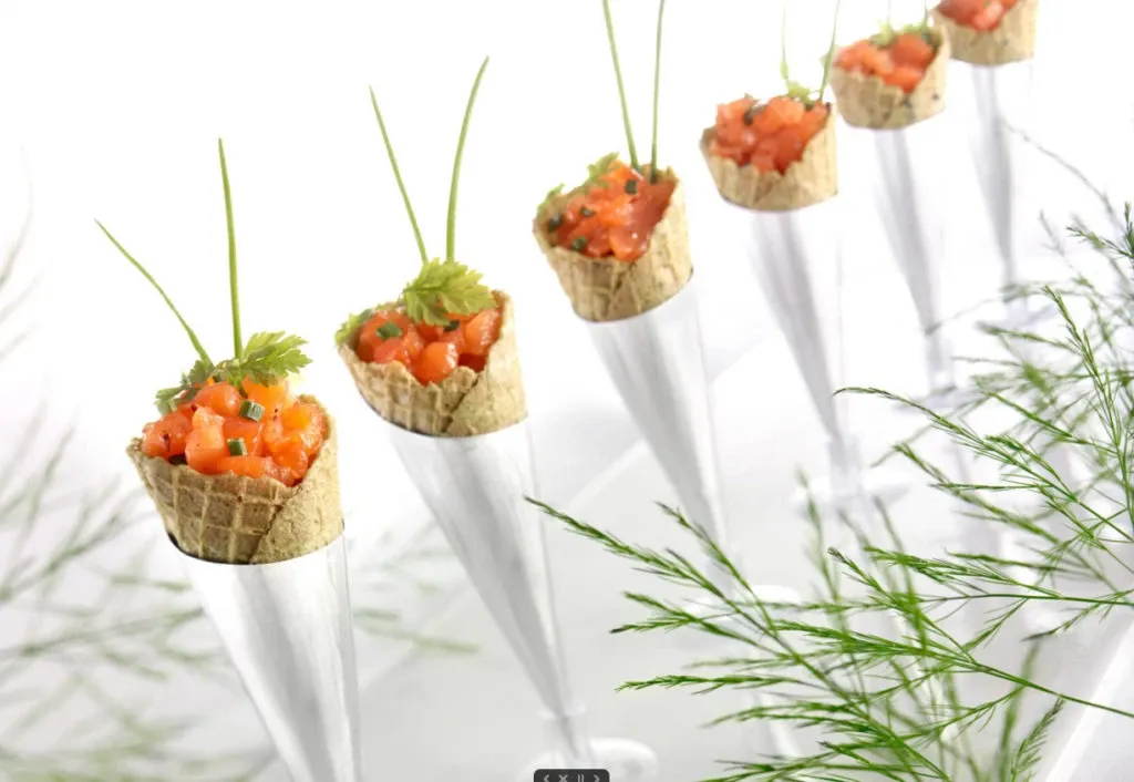 This image showcases a sophisticated appetizer presentation at La Distinction, a renowned wedding venue in Canada, listed under Mariage Elegant's wedding venues category. The image displays several elegantly arranged canapé cones filled with finely diced salmon tartare, garnished with fresh herbs and chives. The cones are neatly positioned upright in sleek, metallic holders, enhancing the visual appeal of a modern, upscale wedding setting. Delicate greenery adds a touch of natural elegance, complementing the overall aesthetic of the venue's catering offering.