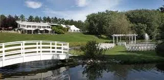 The image showcases La Distinction, a wedding venue featured on Mariage Elegant, Canada. It displays a beautifully landscaped outdoor setting with a white bridge crossing a tranquil pond in the foreground. In the background, a large, elegant building with a spacious patio is visible, complemented by a white tent suitable for receptions. Several rows of white chairs are arranged in front of a charming gazebo, indicating a potential ceremony area. Lush greenery and trees surround the area, offering a serene and picturesque environment for weddings.