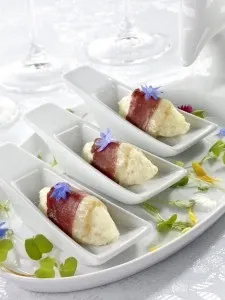 The image shows an elegant food presentation at La Distinction, a wedding venue, showcasing three gourmet appetizers elegantly arranged in individual white ceramic spoons. Each appetizer is wrapped delicately in thin slices of prosciutto and garnished with small blue edible flowers, placed on a white tablecloth surrounded by scattered greenery and flower petals, emphasizing a sophisticated and refined atmosphere suitable for a wedding reception.