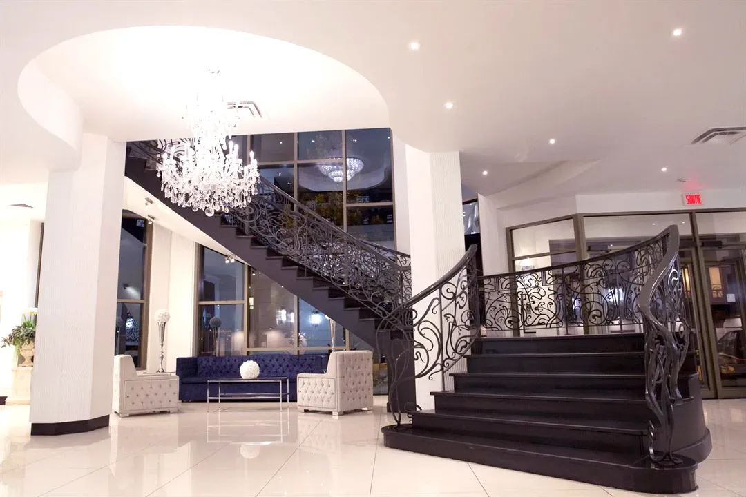 The image showcases the elegant interior of Roma Receptions, a top-tier wedding venue in Canada. The space features a grand, curving staircase with intricate wrought iron railings leading to an upper level. A large, sparkling chandelier hangs from the ceiling, illuminating the entryway with a warm glow. On either side of the staircase, there are plush, tufted white chairs and a sleek, dark blue sofa, offering a stylish and comfortable seating area. The glossy white floor tiles and floor-to-ceiling windows add to the sophisticated ambiance, making Roma Receptions an ideal setting for memorable wedding celebrations.