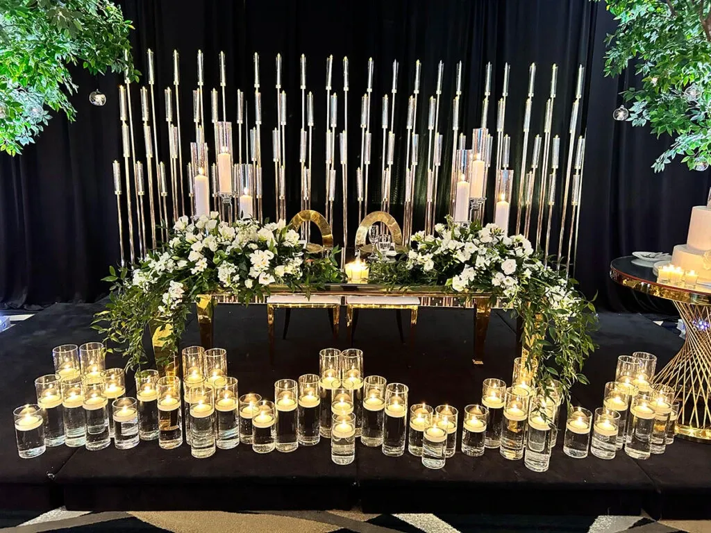 The image showcases a stunning wedding venue setup by Roma Receptions, categorized under Wedding Venues at Mariage Elegant, Canada. The scene features a luxurious arrangement with a central table adorned with an elegant display of white and green floral arrangements. Around the table, numerous floating candles are placed in sleek glass vases, creating a romantic and sophisticated ambiance. Behind the table, a backdrop of vertical metallic rods adds a modern touch to the setting, complemented by soft, ambient lighting. This exquisite setup exemplifies a perfect blend of elegance and opulence, ideal for an unforgettable wedding celebration.