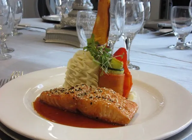 The image features an elegantly arranged dish presented by Roma Receptions, a wedding venue listed under Mariage Elegant, Canada. It showcases a piece of seared salmon garnished with sesame seeds, resting on a vibrant red sauce. Accompanying the salmon is a swirl of creamy mashed potatoes and an assortment of colorful, fresh vegetables, including a rolled cucumber slice, a slice of carrot, and a delicately placed edible garnish on top. The table is set with sparkling wine glasses and silver cutlery, reflecting the sophisticated ambiance of a wedding reception setting. This delightful presentation highlights the culinary offerings at Roma Receptions.