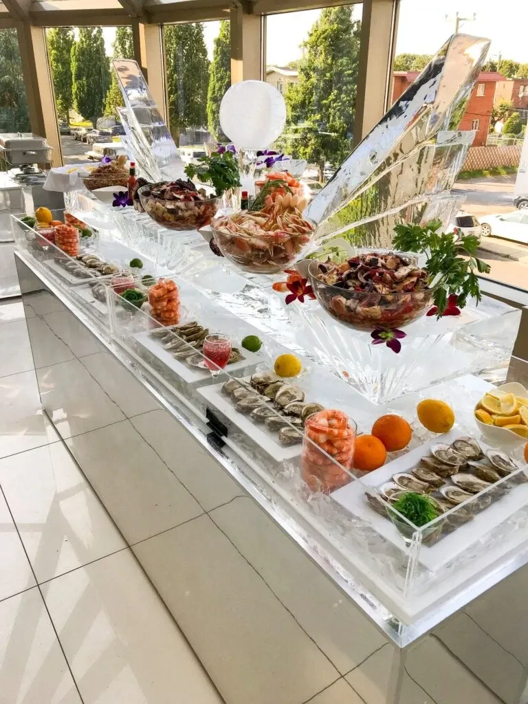 The image showcases an elegant buffet setup at Roma Receptions, a premier wedding venue in Canada. The table is adorned with opulent ice sculptures, each supporting large crystal bowls filled with a variety of fresh salads and garnished with vibrant flowers. Plates of seafood and an array of fresh fruits, including oranges and lemons, are artfully arranged around the sculptures. The background reveals tall windows with views of lush greenery and adjacent buildings, providing a sophisticated and scenic atmosphere perfect for wedding celebrations.