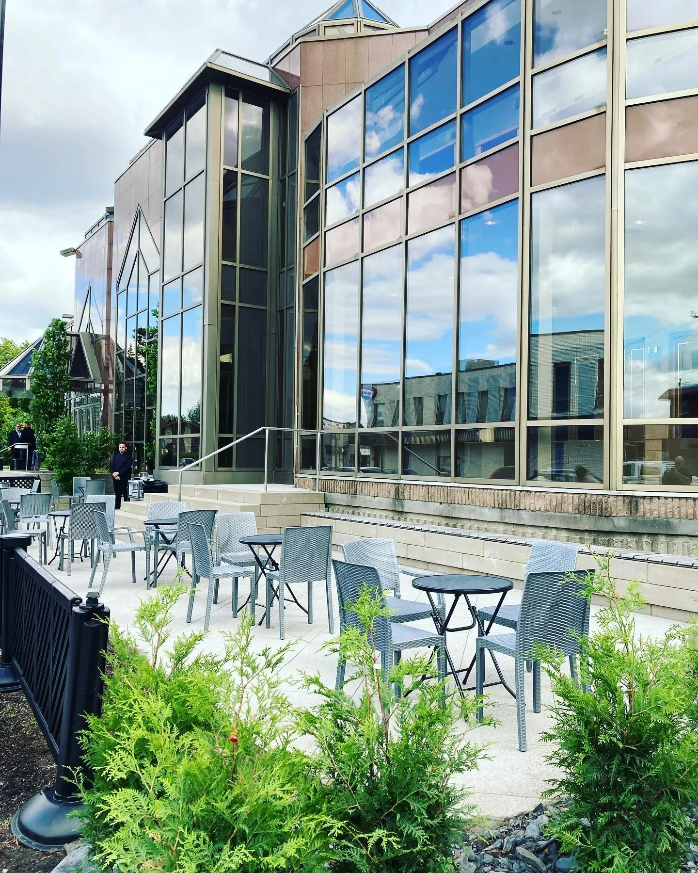 The image shows the elegant exterior of Roma Receptions, a premier wedding venue in Canada, listed on Mariage Elegant. The building features a modern design with large, reflective glass windows that create a grand facade. In the foreground, there's a well-maintained outdoor patio area with several tables and chairs, perfect for guests to enjoy during wedding receptions. The surrounding greenery adds a touch of nature to the refined setting, enhancing the overall ambiance.