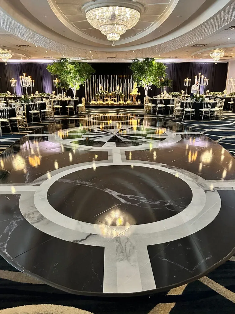 The image showcases an elegant wedding venue by Roma Receptions, featured in the Wedding Venues category. The venue includes a spacious, opulently decorated banquet hall with a glossy black marble dance floor, intricately designed with white circular patterns. The ceiling is adorned with large, luxurious chandeliers that provide a warm and inviting glow. The tables are elegantly set with gold-accented chairs and tall candelabras, enhancing the sophisticated atmosphere. Lush greenery and floral arrangements add a touch of natural beauty, making it an ideal setting for a grand wedding celebration.