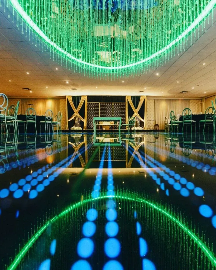 The image showcases a beautifully decorated indoor wedding venue at Roma Receptions, part of the wedding venue listings on Mariage Elegant, Canada. The elegant space features a dazzling ceiling adorned with hanging green lights, creating a stunning visual effect. The floor reflects the intricate light pattern, adding to the ambiance. The venue is set up with modern, transparent chairs arranged neatly around tables, framed by luxurious drapery on the walls, offering a sophisticated setting for weddings.
