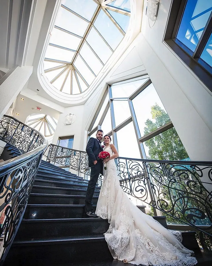 A bride and groom stand gracefully on an elegant, winding staircase at Roma Receptions, a premier wedding venue in Canada. The bride is wearing a stunning, lace-embellished white gown, while the groom is dressed in a classic black suit. They are holding a bouquet of red roses. The staircase features intricate black wrought-iron railings, and the space is filled with natural light streaming in from large, arched windows and a glass ceiling. This sophisticated setting at Roma Receptions highlights the venue's appeal for hosting memorable wedding ceremonies.