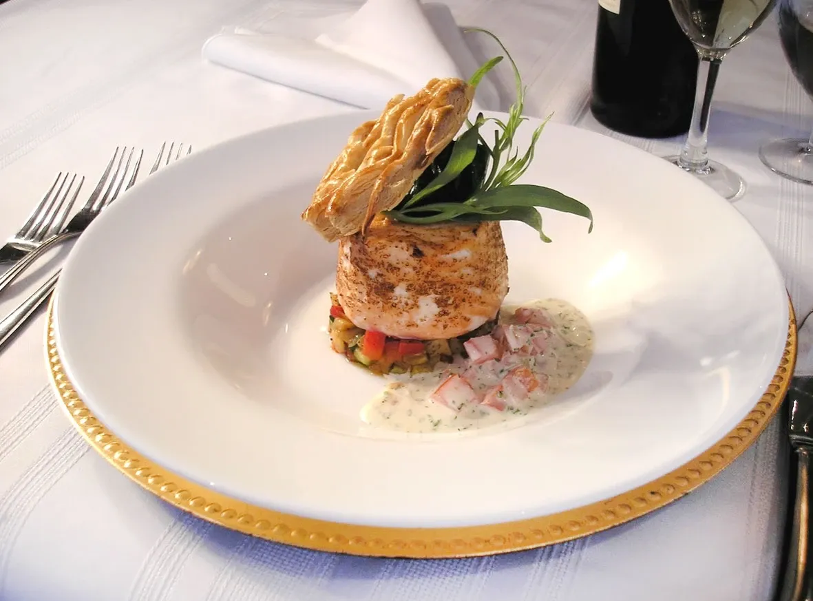 A beautifully plated gourmet dish is elegantly presented on a white plate with a gold-trimmed edge at Roma Receptions, a distinguished wedding venue in Canada. The dish features a seasoned main protein, possibly chicken, artistically garnished with fresh greens and a decorative pastry on top. It is accompanied by a colorful medley of diced vegetables and a creamy sauce, adding to the culinary sophistication. The setting includes neatly arranged silverware and a crisp white tablecloth, suggesting a refined dining experience suitable for wedding receptions.