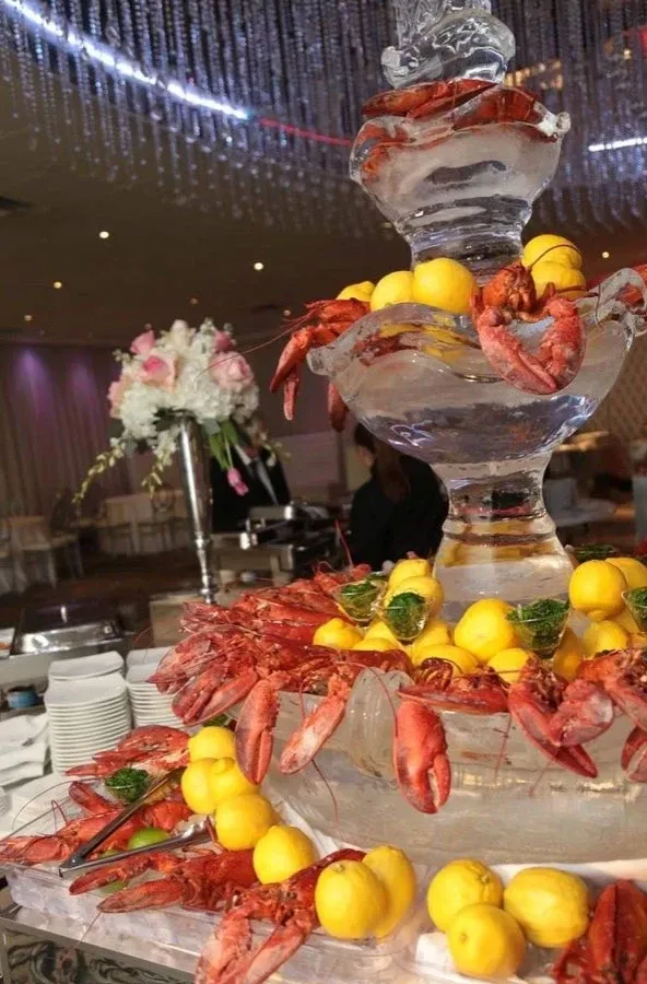 The image showcases an elegant seafood display at Roma Receptions, a top-tier wedding venue under the Wedding Venues category for Mariage Elegant in Canada. It features a grand, multi-tiered ice sculpture centerpiece adorned with vibrant red lobsters and bright yellow lemons. Fresh white plates are neatly arranged to the side, ready for guests. In the background, delicate white floral arrangements add a romantic touch, enhancing the luxurious setting of the venue. The atmosphere is further elevated by the soft glow of purple lighting and a sparkling ceiling decoration, creating a sophisticated ambiance for any wedding celebration.