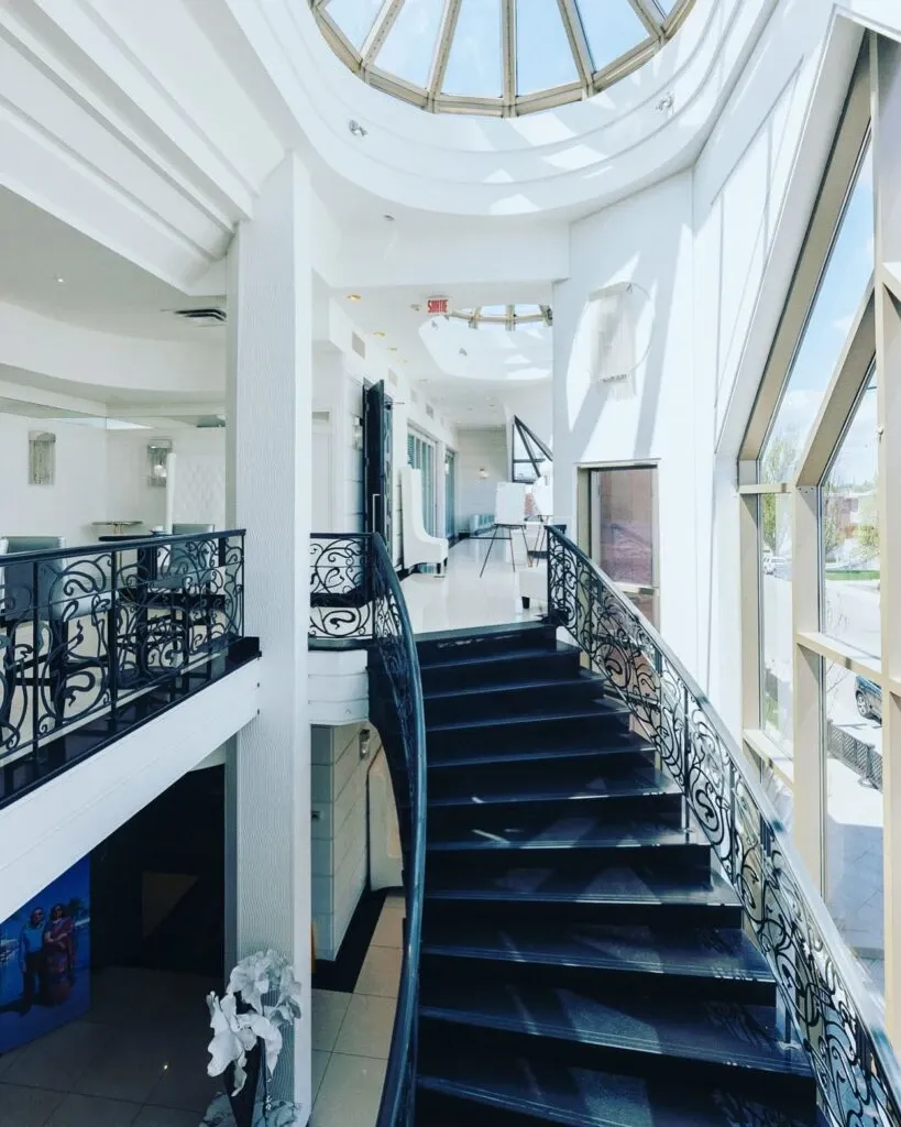 The image showcases the elegant interior of Roma Receptions, a premier wedding venue featured on Mariage Elegant, Canada. It highlights a beautifully designed, curved black staircase with intricate wrought iron railings, leading to an upper-level balcony. The space is illuminated by natural light streaming through large, expansive windows and a skylight, creating a sophisticated and airy atmosphere. The neutral color palette and modern architectural elements make this venue ideal for a stylish and contemporary wedding celebration.