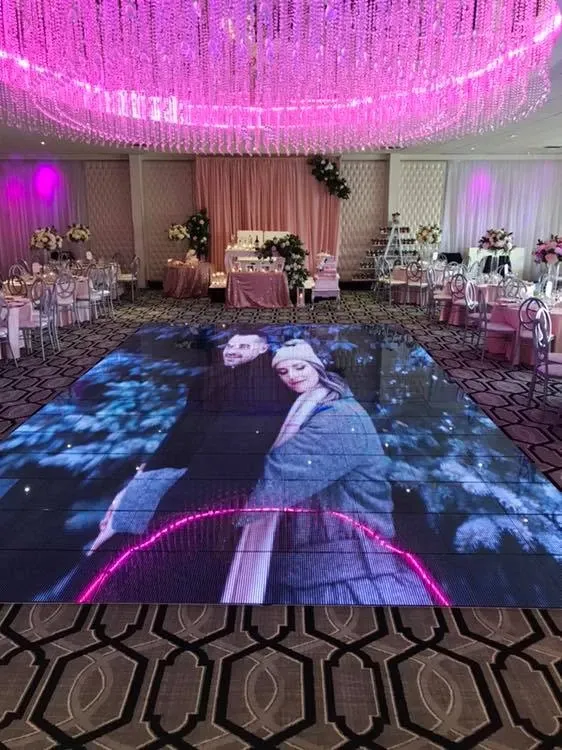 The image showcases the interior of Roma Receptions, a wedding venue under the category of Wedding Venues, featured on Mariage Elegant, Canada. The reception hall is elegantly decorated with a large, illuminated dance floor displaying a romantic image of a couple in a close embrace. Overhead, a stunning pink chandelier composed of cascading lights adds a romantic ambiance to the room. The venue is adorned with tastefully arranged tables, each elegantly set and surrounded by stylish chairs. In the background, pink drapery and lush floral arrangements contribute to the sophisticated and festive atmosphere, perfect for a wedding celebration.