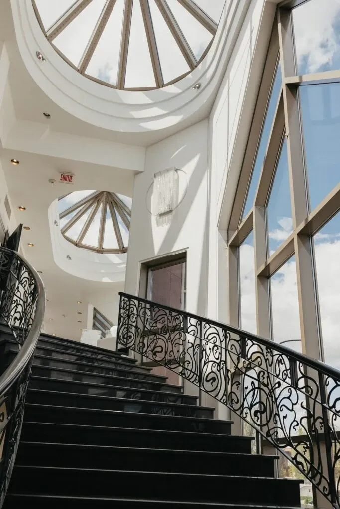 The image showcases the elegant interior of Roma Receptions, categorized under Wedding Venues on Mariage Elegant, Canada. It features a grand staircase with intricate wrought iron railings, leading up to a spacious area illuminated by natural light. Large, modern windows provide a view of the sky, enhancing the sophisticated atmosphere. The ceiling is adorned with a stunning skylight dome, creating a bright and inviting space ideal for wedding events.