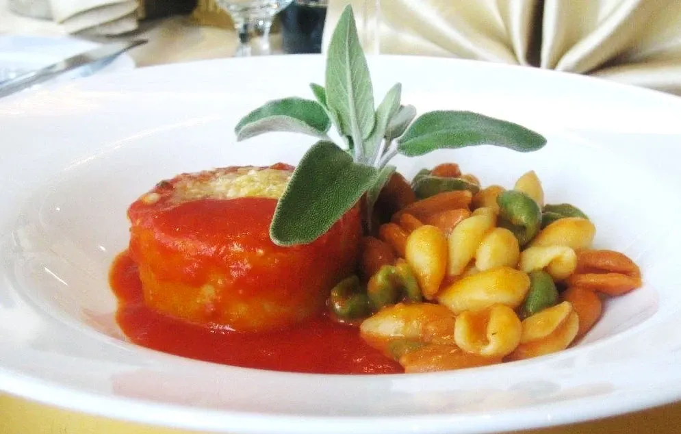 In this image from Roma Receptions, a featured wedding venue listed on Mariage Elegant in Canada, a beautifully presented gourmet dish is displayed. The dish consists of a round portion of layered pasta covered in a rich tomato sauce, garnished elegantly with fresh green sage leaves. Beside it, there is a colorful serving of tri-colored pasta shells, enhancing the visual appeal with hues of green, orange, and white. The sophisticated presentation reflects the venue's commitment to exquisite dining experiences for wedding celebrations.