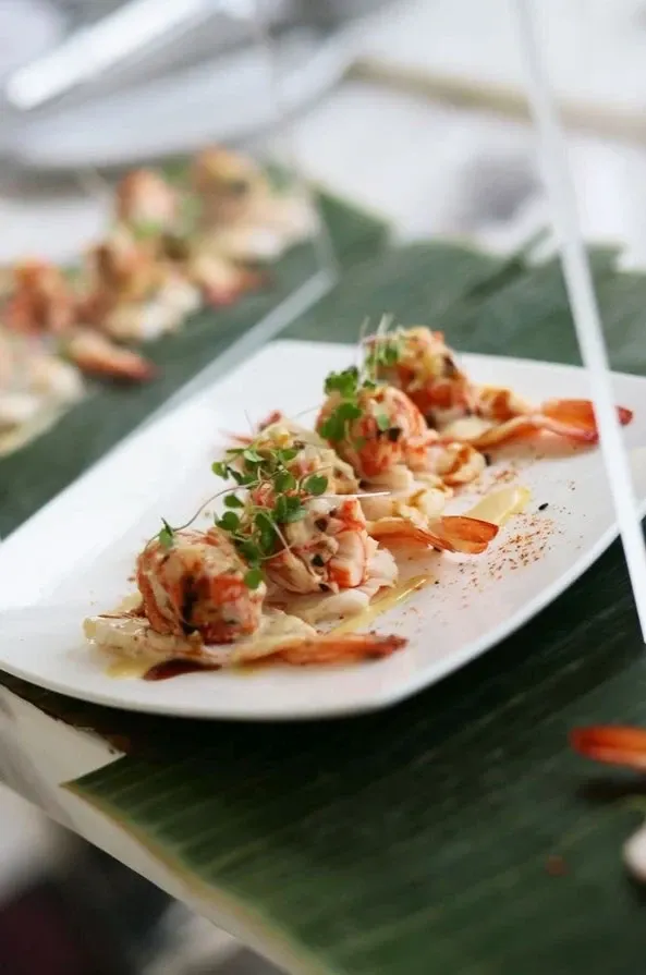 The image shows a beautifully plated seafood dish, featuring shrimp elegantly arranged on a rectangular white plate. The shrimp are garnished with fresh microgreens and drizzled with a creamy sauce. The plate is set on top of large green leaves, adding a natural and tropical touch to the presentation. This culinary presentation is part of the offerings at Roma Receptions, a wedding venue showcased on Mariage Elegant, Canada. The sophisticated dish reflects the high-quality catering expected from a top-tier wedding venue.