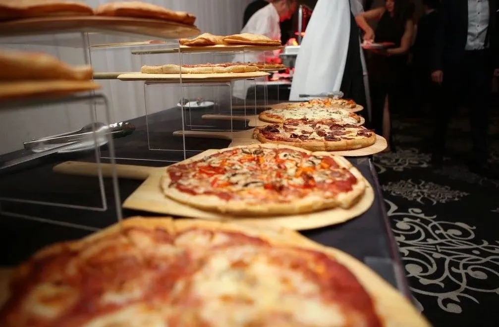 The image shows a wedding reception setup at Roma Receptions, a wedding venue in Canada. In the foreground, a variety of gourmet pizzas are displayed on wooden paddles resting on a modern black table. Each pizza is distinct, featuring toppings such as mushrooms, bell peppers, and various meats, showcasing the venue's diverse catering options. Behind the pizzas, guests are seen mingling in elegant attire, highlighting the venue's suitable ambiance for stylish and relaxed wedding celebrations. The decor features a sophisticated black and white patterned carpet, complementing the elegant setting.