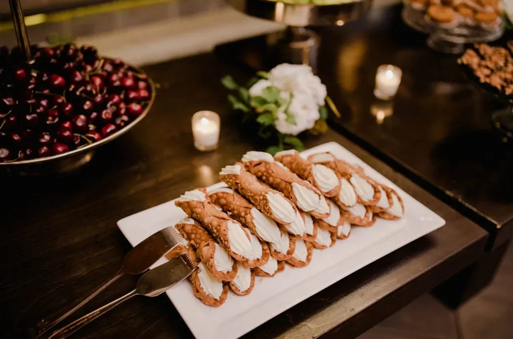 A beautifully arranged display of dessert cannoli on a white platter is present in this image from Roma Receptions, a vendor in the Wedding Venues category. The platter is elegantly positioned on a dark wooden table alongside a bowl of dark cherries and two small, lit candles, adding a romantic ambiance. A white floral bouquet is gently placed behind the platter, enhancing the wedding theme. This setting by Roma Receptions exemplifies the attention to detail and sophistication expected in premier Canadian wedding venues.