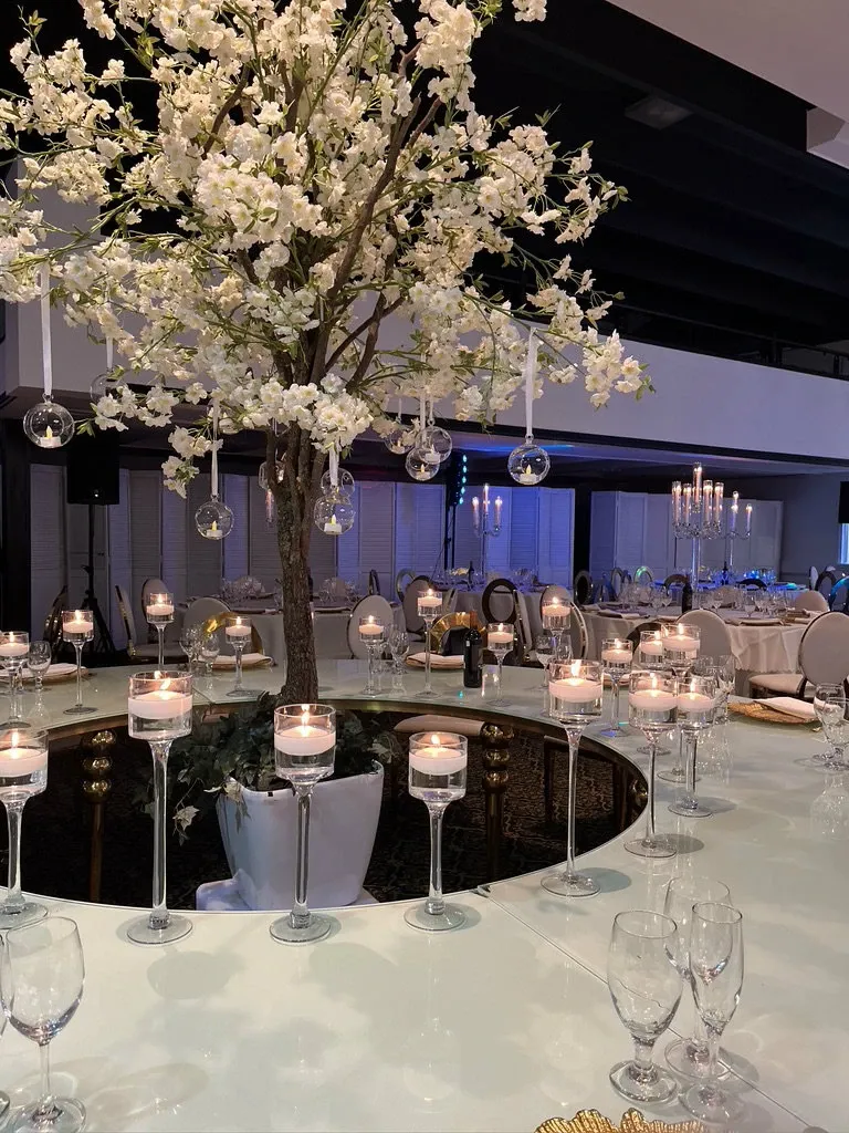 The image showcases the elegant wedding venue, Plaza Le Rizz, a listed vendor under the Wedding Venues category on Mariage Elegant, Canada. The scene features a beautifully decorated round table centerpiece with a tall, white blossom tree adorned with hanging glass orbs. Around the base of the tree, there are numerous tall, lit candles in clear holders, creating a romantic ambiance. The background reveals a sophisticated dining setup with round tables, elegantly arranged chairs, and soft lighting, perfect for a wedding celebration.