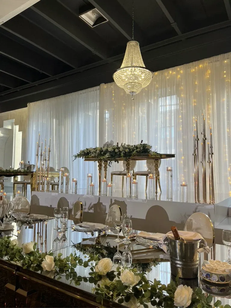 The image showcases an elegantly decorated wedding venue, Plaza Le Rizz, categorized under Wedding Venues for Mariage Elegant in Canada. The setup features a beautifully adorned head table with lush white floral arrangements and greenery running along its edge. Crystal-clear glassware and polished silverware are neatly set, enhancing the luxurious ambiance. A stunning chandelier hangs above, illuminating the area with a warm glow. The backdrop consists of sheer white curtains complemented by twinkling string lights, adding a romantic touch to the venue’s sophisticated atmosphere.