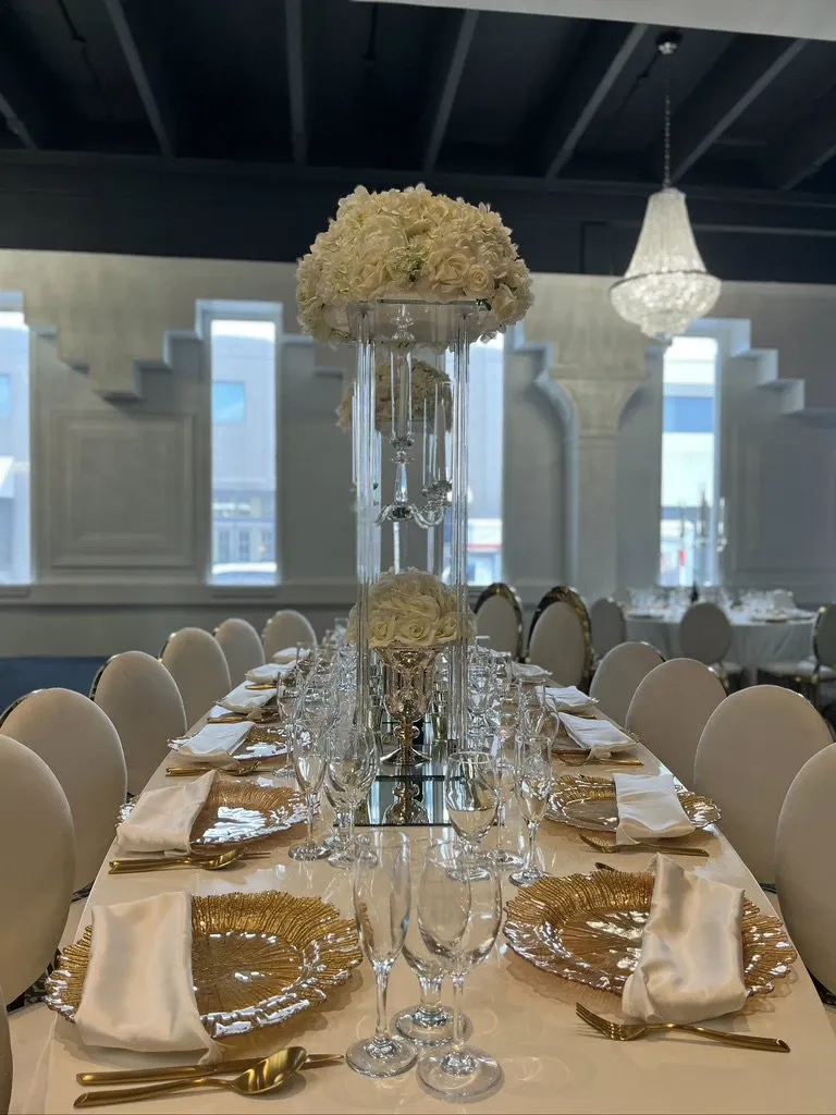 The image showcases an elegant wedding venue table setting at Plaza Le Rizz, categorized under Wedding Venues for Mariage Elegant, Canada. The table is beautifully arranged with a central, towering floral centerpiece featuring white roses in a glass vase. Surrounding the centerpiece are gold-colored decorative plates, neatly folded white napkins, and an array of crystal glassware, all set on a long banquet-style table. The sophisticated ambiance is enhanced by soft natural lighting through large windows and an ornate chandelier hanging from the ceiling, creating a luxurious and inviting atmosphere for wedding celebrations.