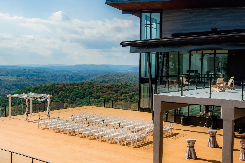 The image showcases La Montagne Coupée, a stunning wedding venue in Canada, under the category of Wedding Venues. It features an elegant outdoor ceremony setup with rows of clear chairs neatly arranged on a spacious wooden terrace, overlooking a breathtaking view of rolling green hills under a partially cloudy sky. A beautifully decorated wedding arch stands at the forefront, while the modern architectural design of the venue's building, with large glass windows, is visible on the right. This serene setting provides a picturesque backdrop ideal for a memorable wedding celebration.