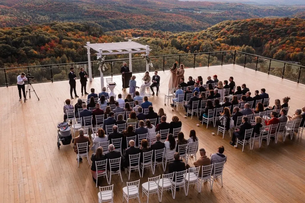 The image showcases a breathtaking wedding venue at La Montagne Coupée, a featured vendor in the Wedding Venues category on Mariage Elegant, Canada. The scene is set on a vast wooden deck overlooking a stunning landscape of rolling hills adorned with autumn foliage. White chairs are neatly arranged in rows facing a charming white pergola, under which the wedding ceremony is taking place. Guests are seated, attentively watching the proceedings, while two photographers capture the moment. The expansive view of colorful trees and distant mountains creates a serene and romantic backdrop, perfect for a memorable outdoor wedding.